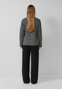 Persoon met lang blond haar, gekleed in een grijze ribbeltrui en zwarte wijde broek, staande tegen een effen lichtgrijze achtergrond.