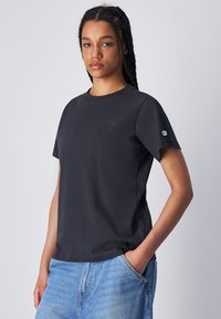Zwarte katoenen t-shirt met korte mouwen, een ronde hals en een klein geborduurd detail op de borst, gecombineerd met lichtblauwe denim jeans.
