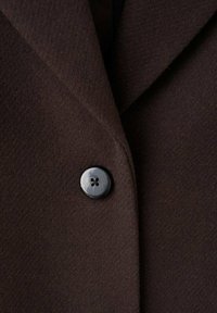 Bruine wollen jas met een gestructureerde afwerking. Voorzien van een gerichette kraag en een zwarte knoop met vier gaten, geplaatst aan de linkerkant.