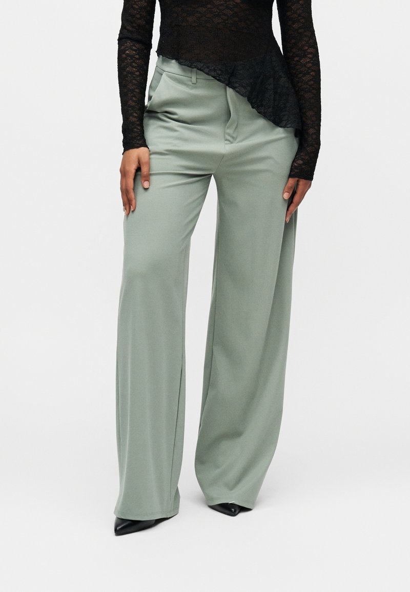 Femme portant un pantalon large taille haute vert clair associé à un haut noir en dentelle à manches longues et des chaussures noires pointues.