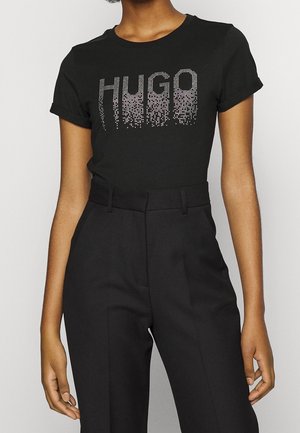 Osoba nosząca dopasowany czarny t-shirt z srebrnym napisem "HUGO" w pikselowym stylu oraz czarne spodnie z wysokim stanem.