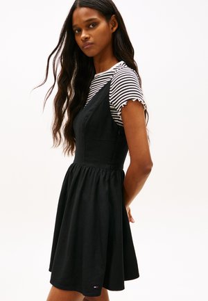 Ung kvinde med langt, bølget hår iført en sortærmeløs kjole over en sort-hvid stribet kortærmet skjorte, stående sidelæns.