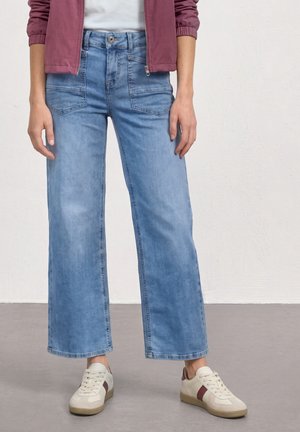 Persoon die lichtblauwe wijde jeans draagt, gebroken witte sneakers met bruine strepen, een wit overhemd en een bordeauxrode jas met opgerolde mouwen.