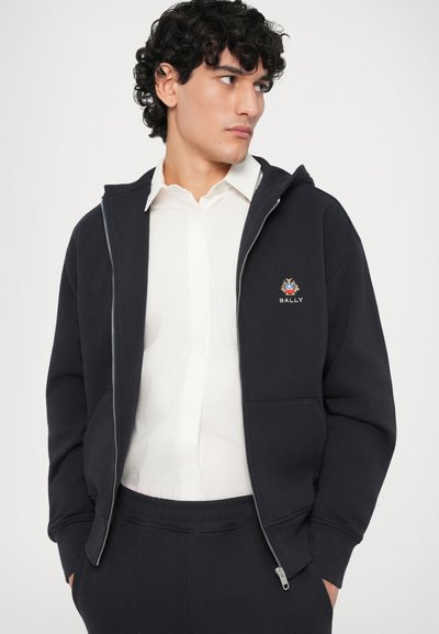 Sudadera con cremallera en color azul marino, fabricada en tela suave, con un bolsillo frontal y el logo de Bally bordado. Combinada con una camiseta blanca para crear contraste.