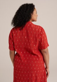 Blusa corta roja de manga corta con una tela texturizada. Presenta rayas diagonales metálicas doradas y un cuello tipo camisa. Se drapea de manera suelta.
