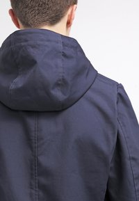 Giacca navale con cappuccio, caratterizzata da una texture liscia e un design semplice. Cuciture visibili lungo le spalle e la schiena.