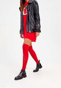 Chaqueta de cuero negra de motociclista sobre un vestido rojo, combinada con calcetas rojas hasta el muslo y botas negras brillantes con cremallera lateral.
