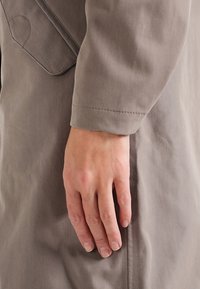 Capo spalla grigio con una texture liscia; caratterizzato da una manica sagomata e un dettaglio discreto della tasca. Mano appoggiata sul capo.