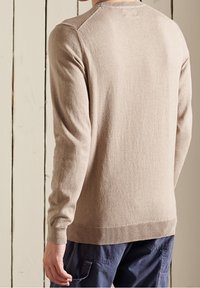 Pull beige en matière douce, avec manches longues, poignets côtelés et col rond classique. Texture visible et style simple.