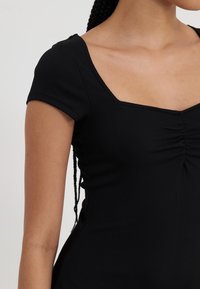 Černý žebrovaný top s krátkými rukávy, čtvercovým výstřihem a nařasenými detaily vpředu. Měkká, elastická tkanina s přiléhavým střihem.