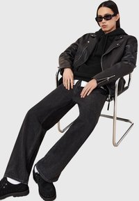 Veste de moto en cuir noir par-dessus un sweat à capuche noir, associée à un jean large noir et des chaussures noires. Des lunettes de soleil élégantes et des bagues complètent le look.
