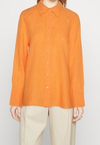Camicia di lino arancione con colletto a punta, maniche lunghe, chiusura a bottoni e vestibilità rilassata. Tessuto testurizzato con una leggera lucentezza.