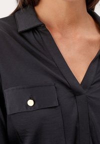 Camicia nera con colletto a V, dotata di tasca con un bottone dorato come dettaglio, texture liscia. Focus su un design minimalista e linee pulite.