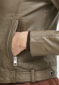 Olivovo zelená kožená bunda s vreckom na zips, ribovanými manžetami a detailom opasku v oblasti pása. Hladká textura s jemným leskom.