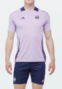 Lila atletisk tröja med mörkblå detaljer, prydd med en tupp-emblem och Adidas-logotyp. Kombinerad med marinblå shorts som har en liknande tuppdesign.