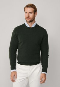 Maglione verde scuro realizzato in morbido tessuto a maglia, con scollatura rotonda e maniche lunghe, abbinato a chinos di colore chiaro.