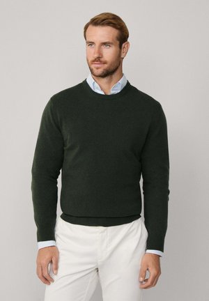 Dunkelgrüner Pullover aus weichem Strickmaterial, mit Rundhalsausschnitt und langen Ärmeln, kombiniert mit hellfarbigen Chinos.