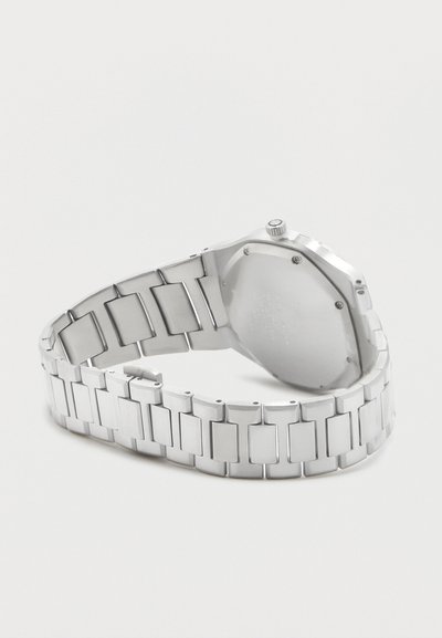 Ασημένιο μεταλλικό ρολόι χειρός που εμφανίζεται από πίσω, αναδεικνύοντας το συνδεδεμένο μπρασελέ και την κλειστή θήκη του ρολογιού με κορώνα στο πλάι.