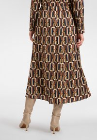 Vestido midi marrón impreso con un patrón de cadena geométrica. El material parece suave; cuenta con una cintura elástica y mangas largas, combinado con botas de caña alta color beige.