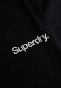 Zwarte fleecetrekker met een V-hals ontwerp. Gekenmerkt door een textuurvol wit "Superdry." logo dat prominent op de voorkant is weergegeven. Zacht materiaal.
