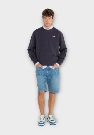 Sweat-shirt bleu marine à col rond avec deux petits points rouges, associé à un short en denim bleu clair et des baskets blanches avec des accents bleus.