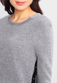 Femme portant un pull en maille gris clair avec un col rond et des détails de boutons noirs sur le côté.