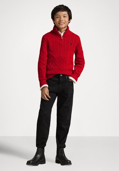 Červený sveter s pleteným vzorom a zipsovým golierom, skombinovaný s čiernymi nohavicami s rovnými nohavicami a čiernymi členkovými topánkami. Jednoduchý, klasický dizajn.