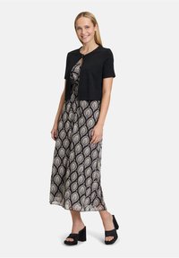 Schwarze, kurzärmlige, cropped Strickjacke über einem gemusterten Maxikleid mit einem Blättermuster in beigen und dunkelblauen Farbtönen. Schwarze Plateauschuhe.