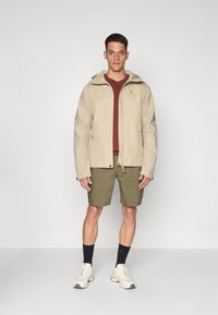 Homme portant une veste à capuche beige sur une chemise bordeaux, un short vert olive, des chaussettes noires et des baskets blanches, se tenant contre un fond uni.