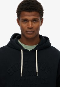 Sudadera navy con cordones color crema y texto en relieve "SUPERDRY N.Y.C. ATHLETICS" en el frente, llevada sobre una camiseta de color verde claro.