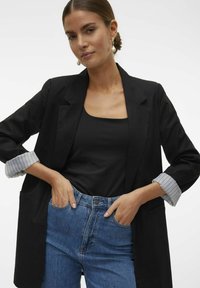 Blazer noir ajusté avec des poignets à rayures blanches, associé à un débardeur noir et un jean bleu taille haute. Design simple et lignes épurées.