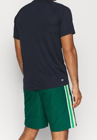 Marineblaues Sporthemd mit kurzen Ärmeln, kombiniert mit grünen Shorts, die weiße und gelbe Seitenstreifen aufweisen. Der Stoff wirkt glatt und leicht.