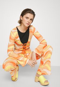 Conjunto de duas peças com um top justo com um decote V profundo e uma fita de renda laranja, feito de um tecido amarelo e laranja com padrões; calças flare a complementar. Tamancos amarelos.