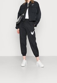 Svart lättviktsjacka med två framfickor, kombinerad med matchande avsmalnande byxor med en vit Nike-logga och vita sneakers.