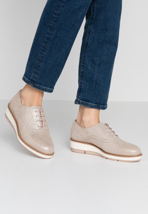 Zapatos oxford de piel beige con diseño de cordones, detalles perforados y una suela gruesa de color claro. Combinados con pantalones de mezclilla oscuros.