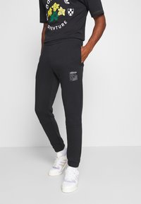 Černé tepláky s elastickými manžetami a bočními kapsami, se záplatou s grafickým designem. V kombinaci s černým tričkem a bílými teniskami.