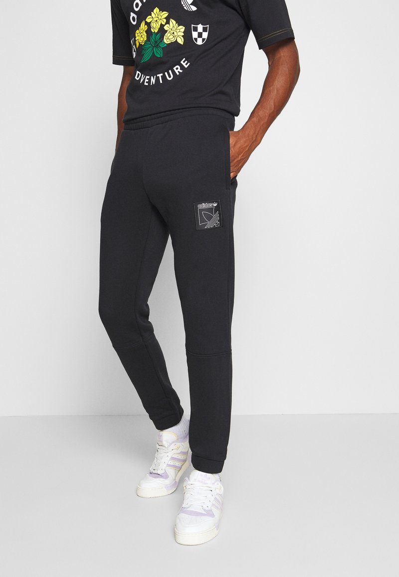 Černé tepláky s elastickými manžetami a bočními kapsami, se záplatou s grafickým designem. V kombinaci s černým tričkem a bílými teniskami.