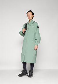 Manteau imperméable vert clair avec une capuche, des poignets élastiques et deux poches latérales. Associé à des bottes de randonnée noires et un legging.