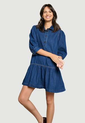 Vrouw glimlacht, draagt een blauwe denimjurk met knopen, hoepeloorbellen en heeft donker haar, staat met één been gekruist voor het andere.