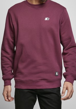 Mężczyzna w prostym bordowym swetrze typu crewneck z małym białym logo na piersi i na dole, w połączeniu z czarnymi spodniami.