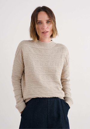 Bež pleteni pulover z teksturiranim zigzag vzorcem, rebrastim ovratnikom in dolgimi rokavi. Uporabljen z temnimi jeans hlačami.