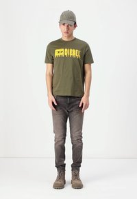 Grønn bomull t-skjorte med gul "DIESEL" trykk, grå caps, grå jeans og beige snøresko. Modell står foran en lys bakgrunn.