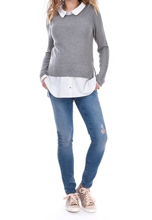 Maglione grigio a maglia con scollo rotondo, indossato sopra una camicia bianca a bottoni. Abbinato a jeans blu strappati e sneakers rosa chiaro.