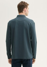 Ciemnozielona koszulka polo z długim rękawem, z kołnierzykiem, gładką teksturą i bocznymi rozcięciami na dole. Brak widocznych wzorów lub detali metalowych.