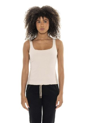 Une femme aux cheveux bouclés volumineux se tient devant un fond blanc. Elle porte un débardeur blanc et un pantalon noir à cordon de serrage.