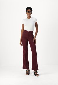 Pantaloni palazzo in velluto a coste bordeaux a vita alta con dettagli in bottone dorato, abbinati a una maglietta attillata bianca a maniche corte con riga e sandali neri con suola spessa.