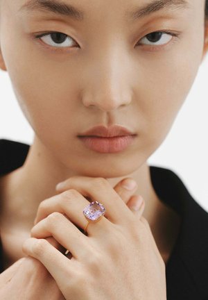 Anillo de oro con una gran piedra preciosa de corte cuadrado en color lavanda. Los tonos de piel son naturales y el fondo es neutro y poco intrusivo.