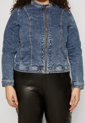 Kobieta w wyblakłej, niebieskiej kurtce dżinsowej z zamkiem błyskawicznym z przodu i czarnych skórzanych spodniach, widoczna od szyi do bioder.