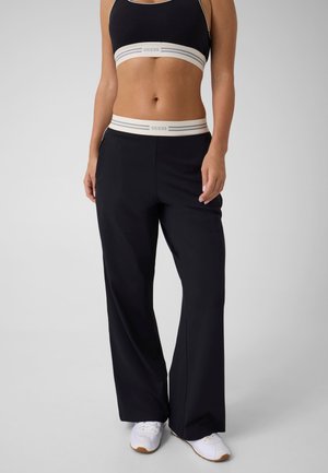 Femme portant un pantalon noir à jambes larges et un soutien-gorge de sport noir avec des bandes élastiques blanches marquées, associé à des baskets blanches sur fond clair.