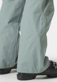 Lichtgroene waterdichte skibroek met een slank ontwerp, voorzien van een logo nabij de knie, en gecombineerd met zwarte sneeuwschoenen.